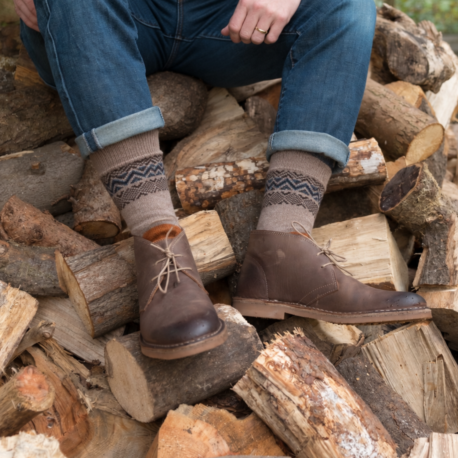 A person sitting on a pile of cut logs wearing brown leather lace-up boots, patterned knit socks, and blue jeans.