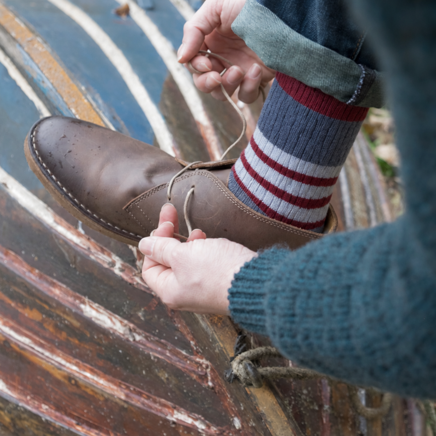 A close-up lifestyle shot of a person tying the laces of a brown leather shoe, wearing a blue knit sweater, striped socks, and blue jeans, set against a rustic wooden background.