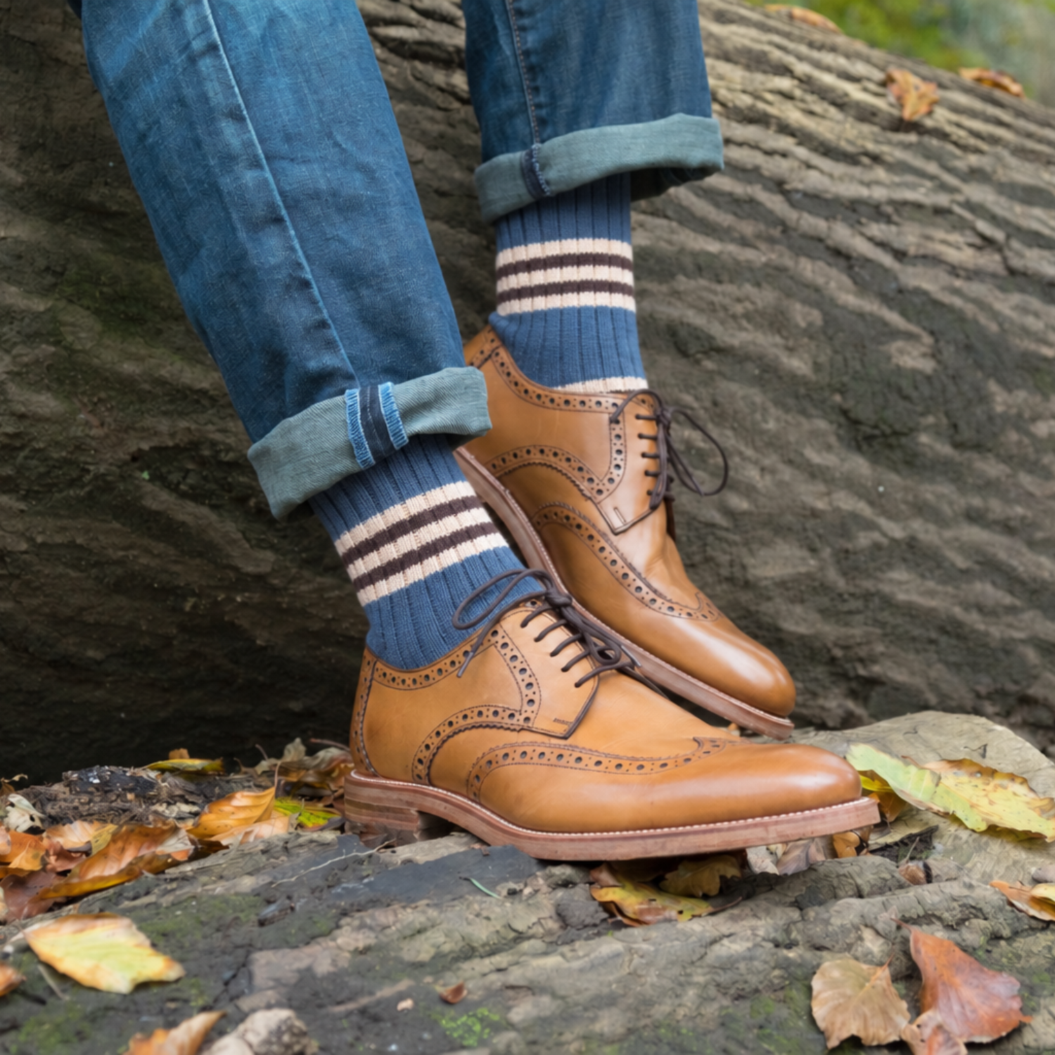 A person wearing blue jeans with cuffed hems, navy blue socks with white stripes, and tan leather wingtip oxford shoes, standing outdoors on a mossy log with scattered autumn leaves.