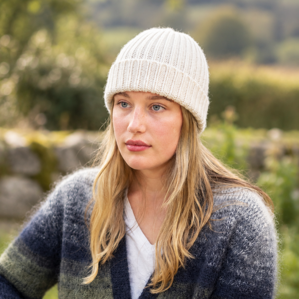 A woman wearing a cream ribbed knit beanie and a blue-gray striped knit cardigan, photographed outdoors with a blurred natural background.