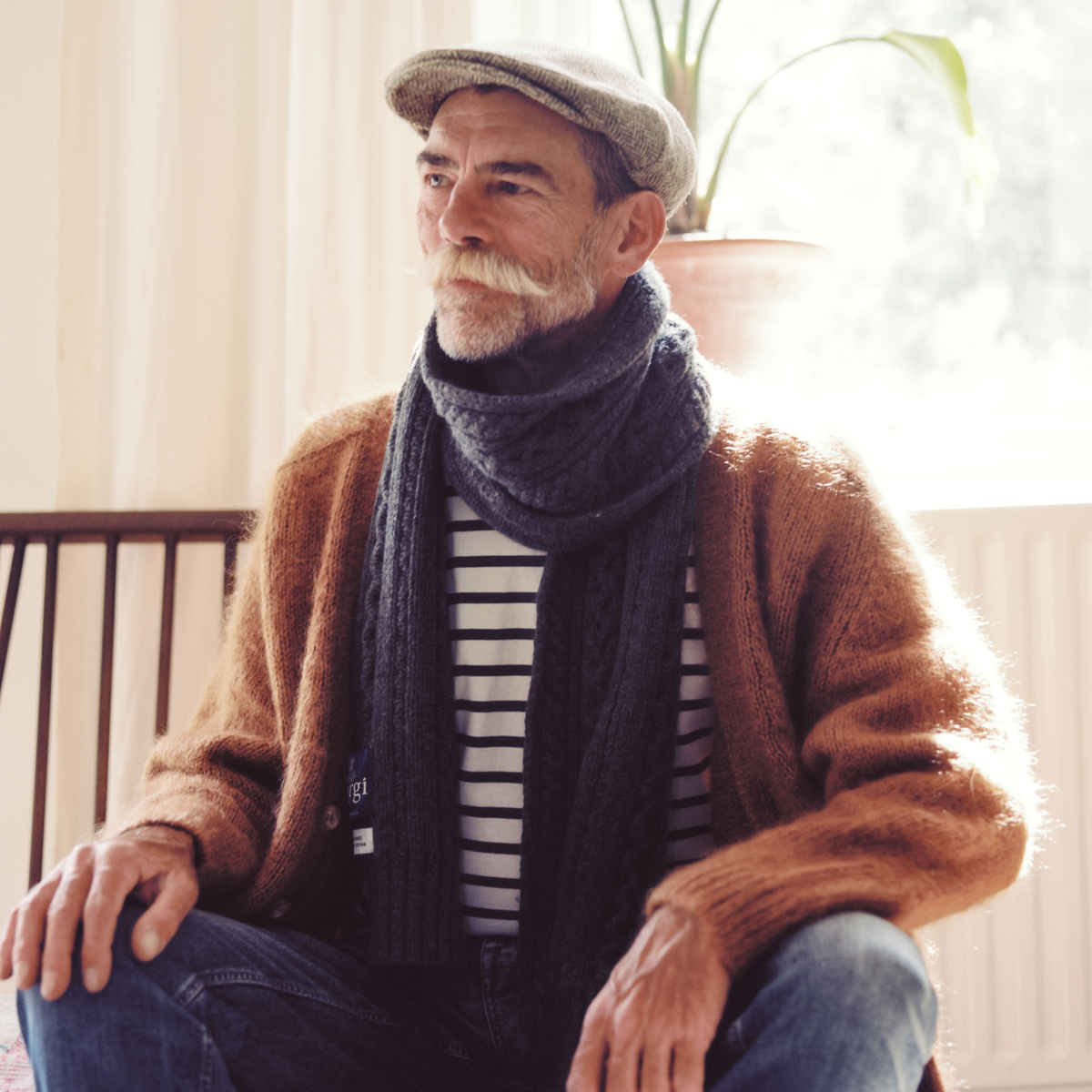 A man with a white mustache wearing a brown knit cardigan, a dark gray cable-knit scarf, a striped shirt, and a flat cap, sitting in a brightly lit indoor setting.