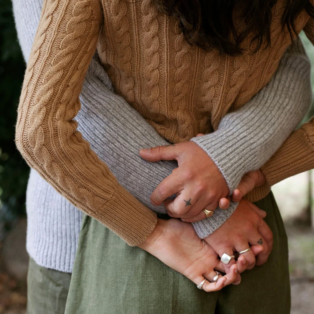 Two people embracing outdoors, with one person wearing a tan cable-knit sweater and olive green skirt, and the other wearing a light gray ribbed sweater. Their hands are clasped together, showing off multiple silver rings.