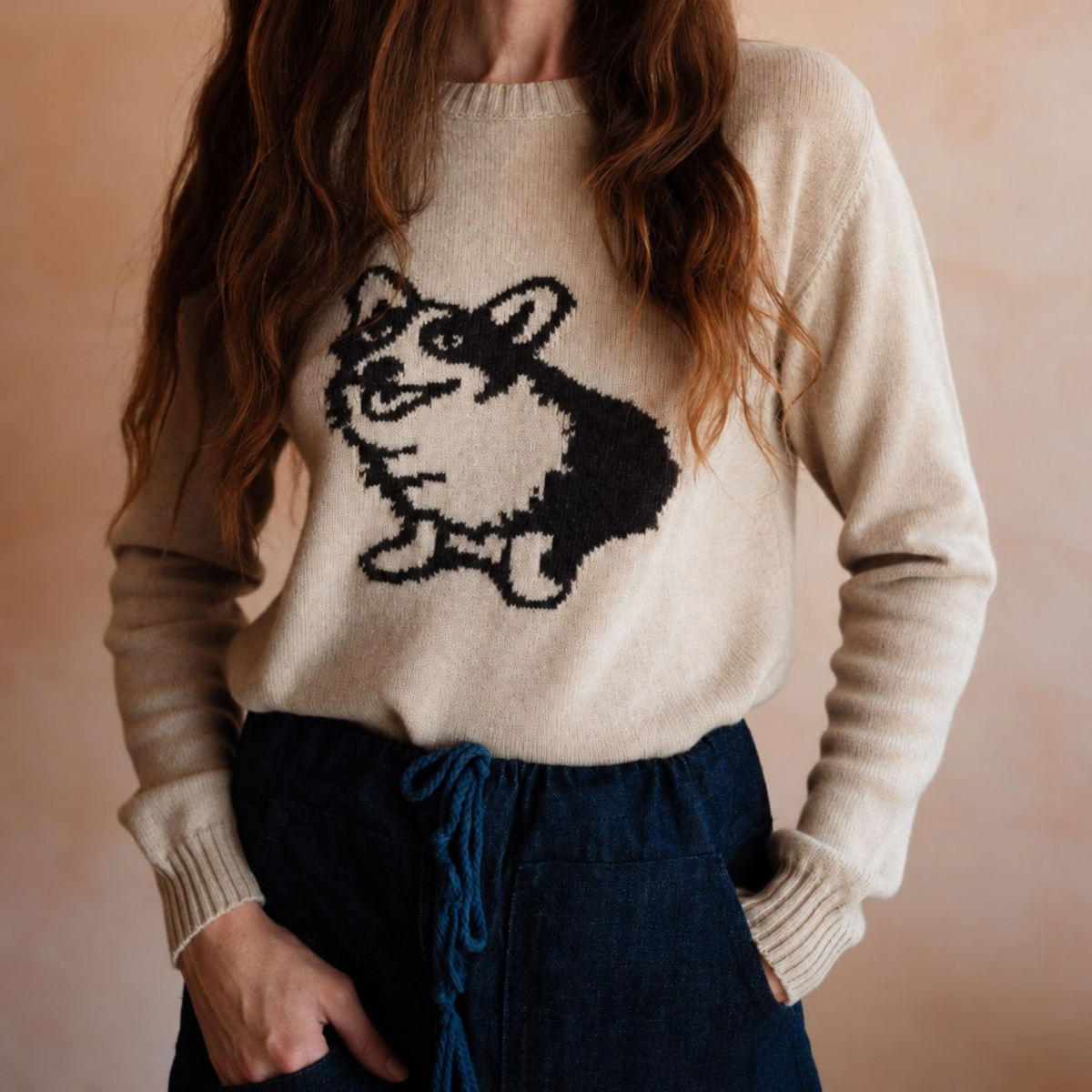 A woman wearing a cream-colored knit sweater with a black and white corgi graphic on the front, paired with dark blue jeans, set against a warm beige background.