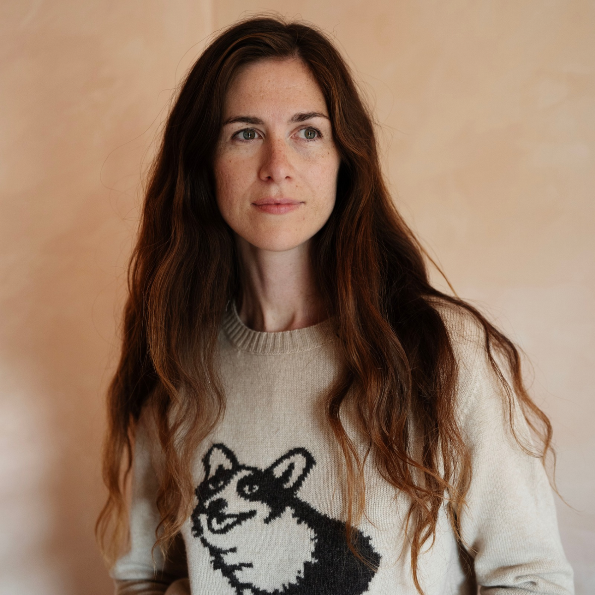 A woman with long wavy brown hair wearing a cream knit sweater with a black dog graphic, photographed against a soft beige background.