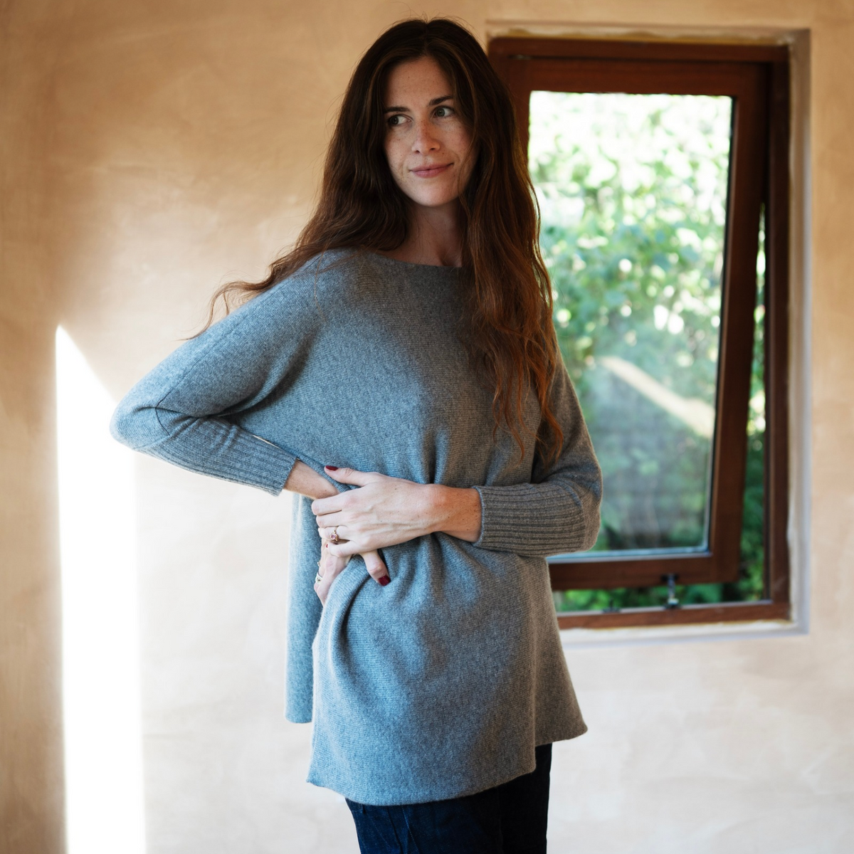 A woman with long brown hair wearing a light blue-gray knit sweater and dark jeans, standing in a warm-toned room with a window behind her.