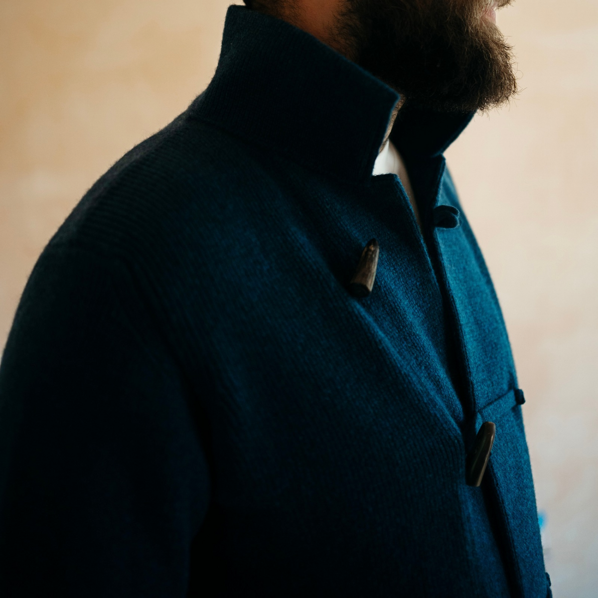 Close-up of a man wearing a dark navy blue textured wool-blend coat with a high collar and dark toggle closures, set against a soft beige background.