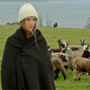 Woman wrapped in a blanket and hat standing in a field with sheep