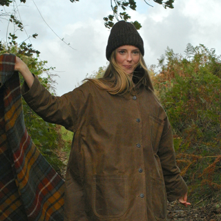 Person wearing a brown coat and chocolate brown beanie style knit hat standing outdoors with greenery in the background