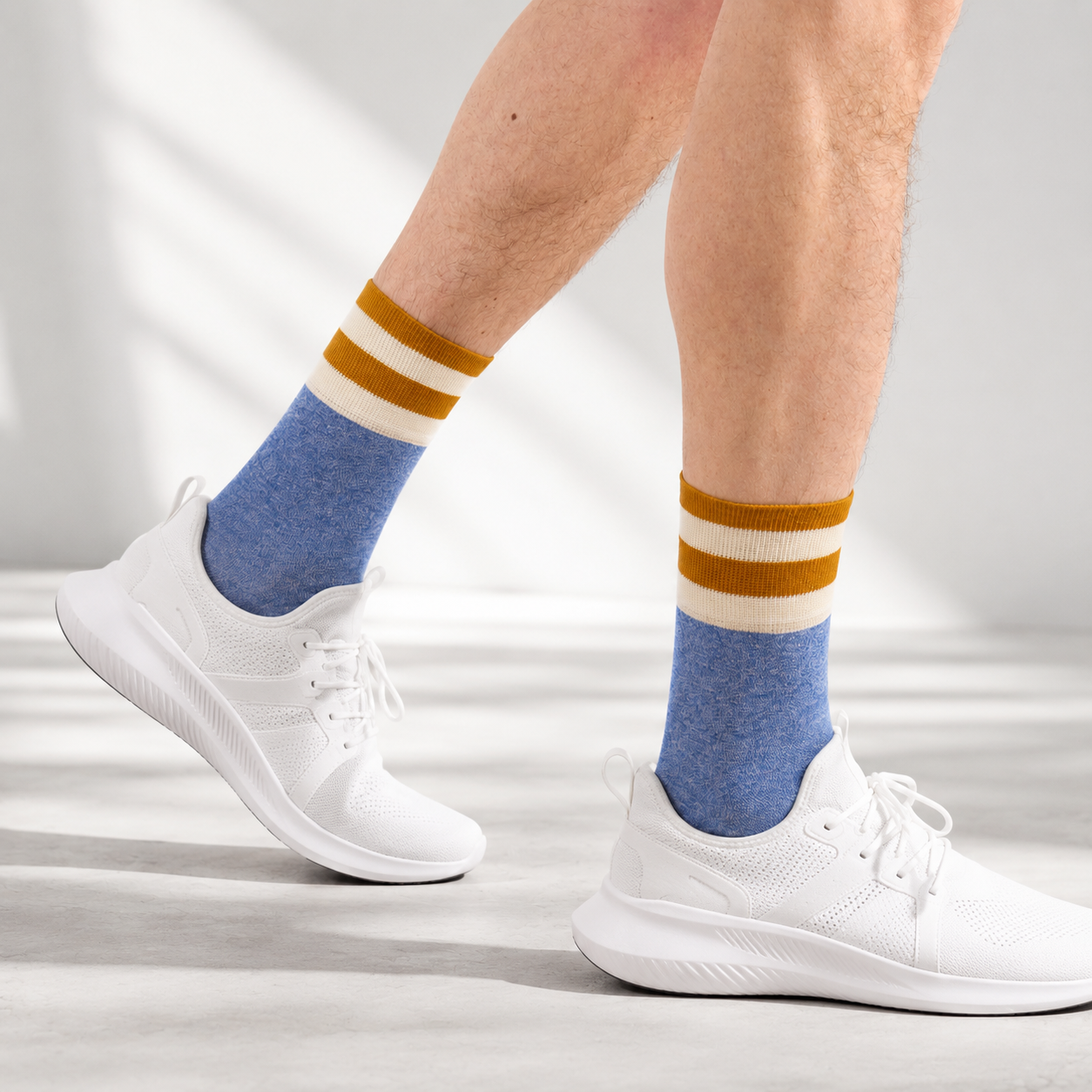 A person's legs wearing heather blue crew socks with two horizontal stripes in mustard yellow and cream, paired with white athletic sneakers on a light gray floor with soft window shadows.