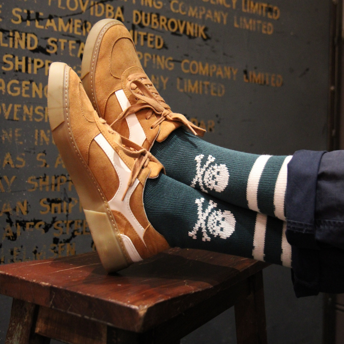 Brown suede shoes and dark green socks with white skull designs on a wooden stool against a textured wall.