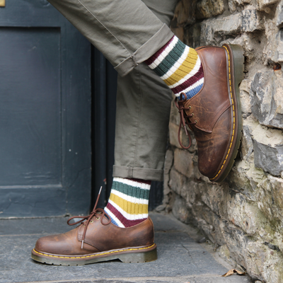 Person wearing brown boots and colorful striped socks against a stone wall.
