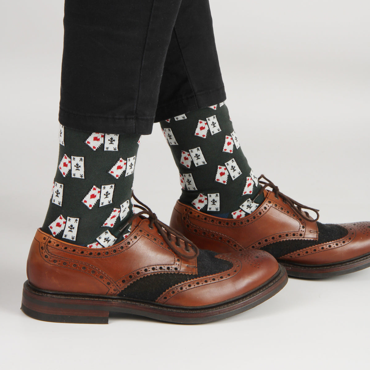 A person wearing black pants, black socks with a repeating playing card pattern, and brown brogue shoes on a light gray studio background.