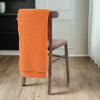 An orange wool blanket with a fine honeycomb texture draped over the back of a wooden chair, set against a background of a simple indoor scene with a dark chair and a white door.