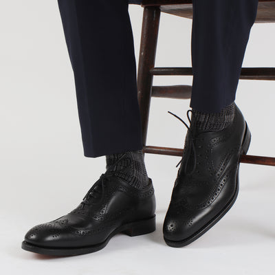 A pair of men's formal woven check cotton socks in black and white, displayed alongside a pair of black dress shoes and navy pants.