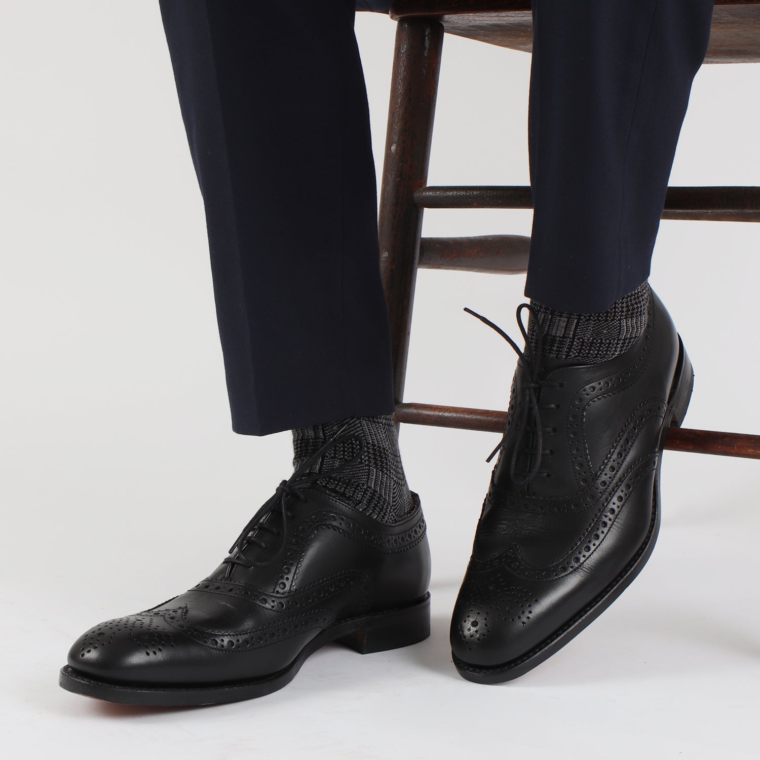 A pair of men's formal woven check cotton socks in black and white, displayed alongside a pair of black dress shoes and navy pants.