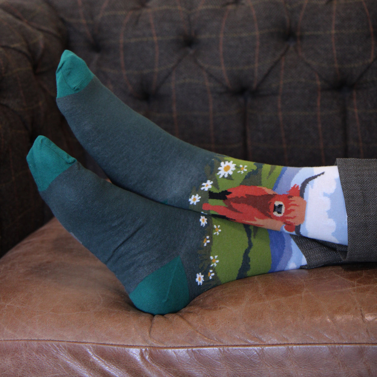 A pair of dark teal crew socks with a colorful graphic of a red cow in a field of white daisies, shown on feet resting on a brown leather couch.