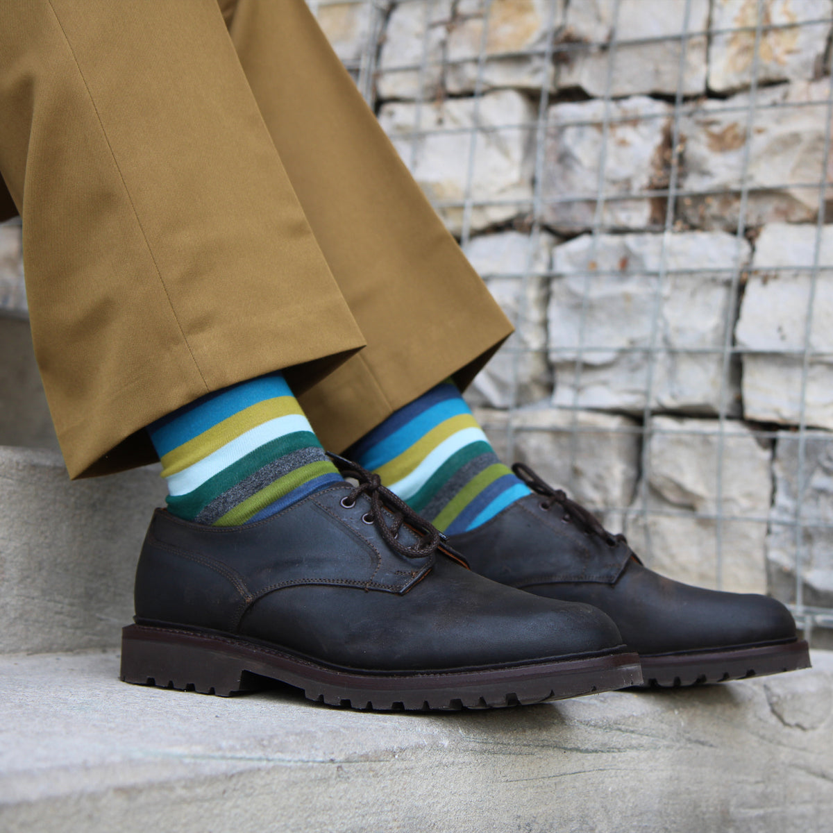 A person seated on stone steps wearing dark brown leather lace-up shoes with colorful striped socks and tan trousers, against a stone wall with a wire grid overlay.