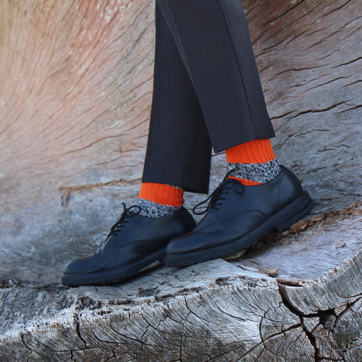 A person wearing black dress shoes and bright orange socks with a gray patterned cuff, standing on a weathered tree stump outdoors.