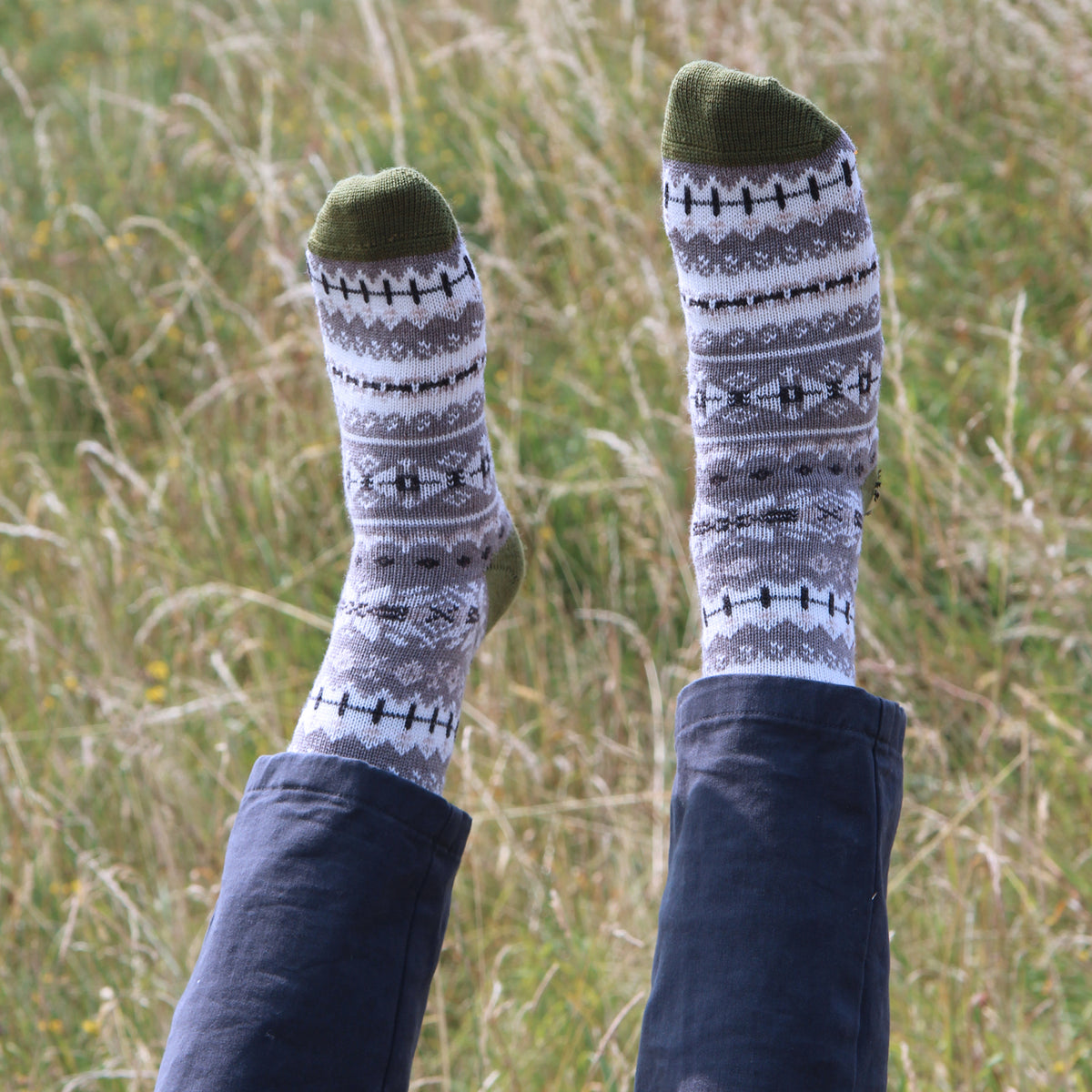 A person lying on grass outdoors with their legs raised, wearing a pair of gray and white patterned knit socks with olive green toes and heels, paired with dark blue jeans.