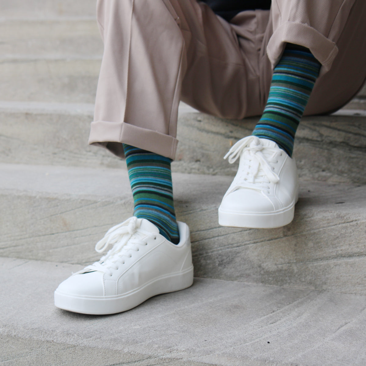 A person sitting on concrete steps wearing white low-top sneakers, beige pants, and striped socks in shades of blue, green, and teal.