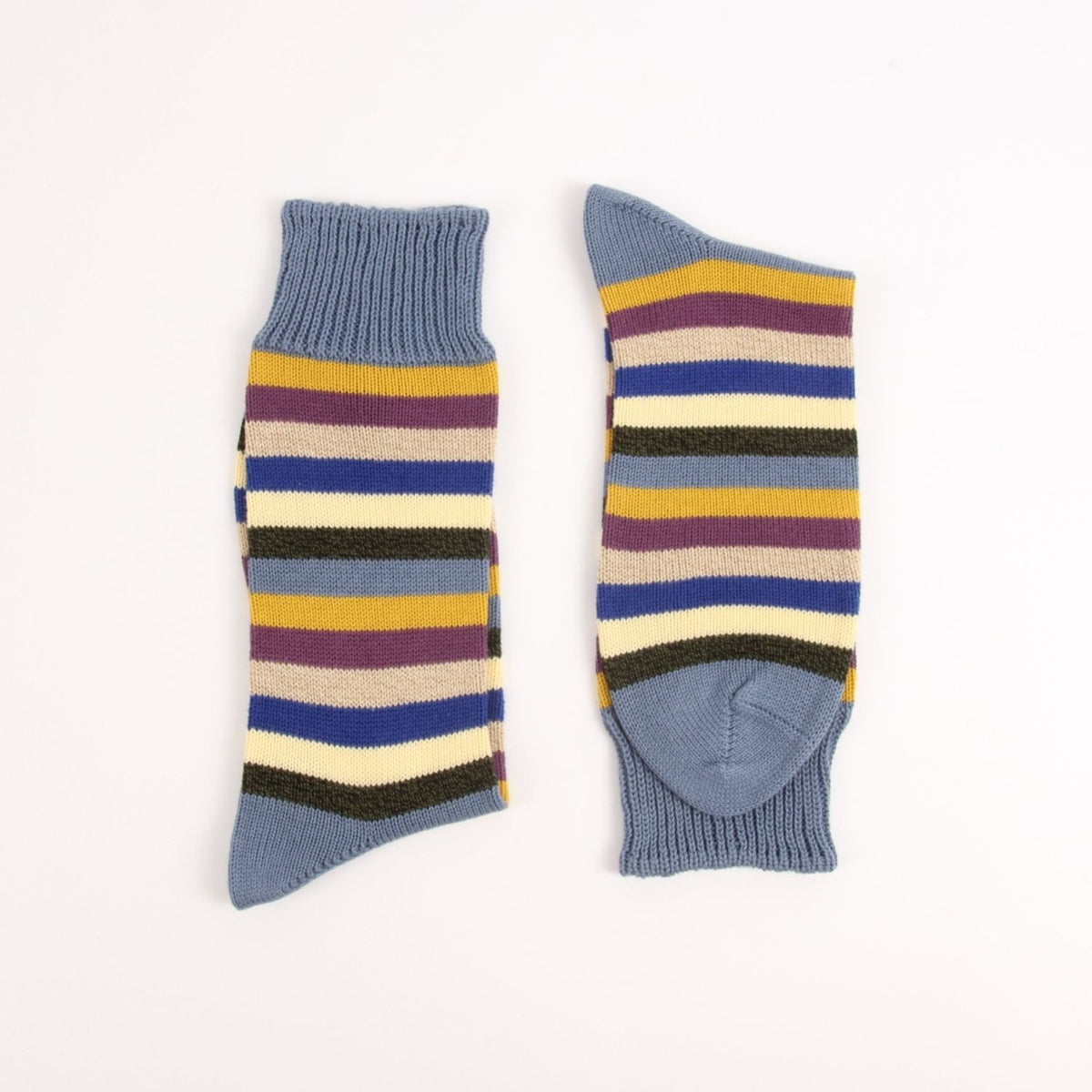 A pair of striped crew socks laid flat on a white background, showing both the front and side views. The socks feature a dusty blue ribbed cuff, toe, and heel, with horizontal stripes in yellow, purple, dark green, cream, and blue.