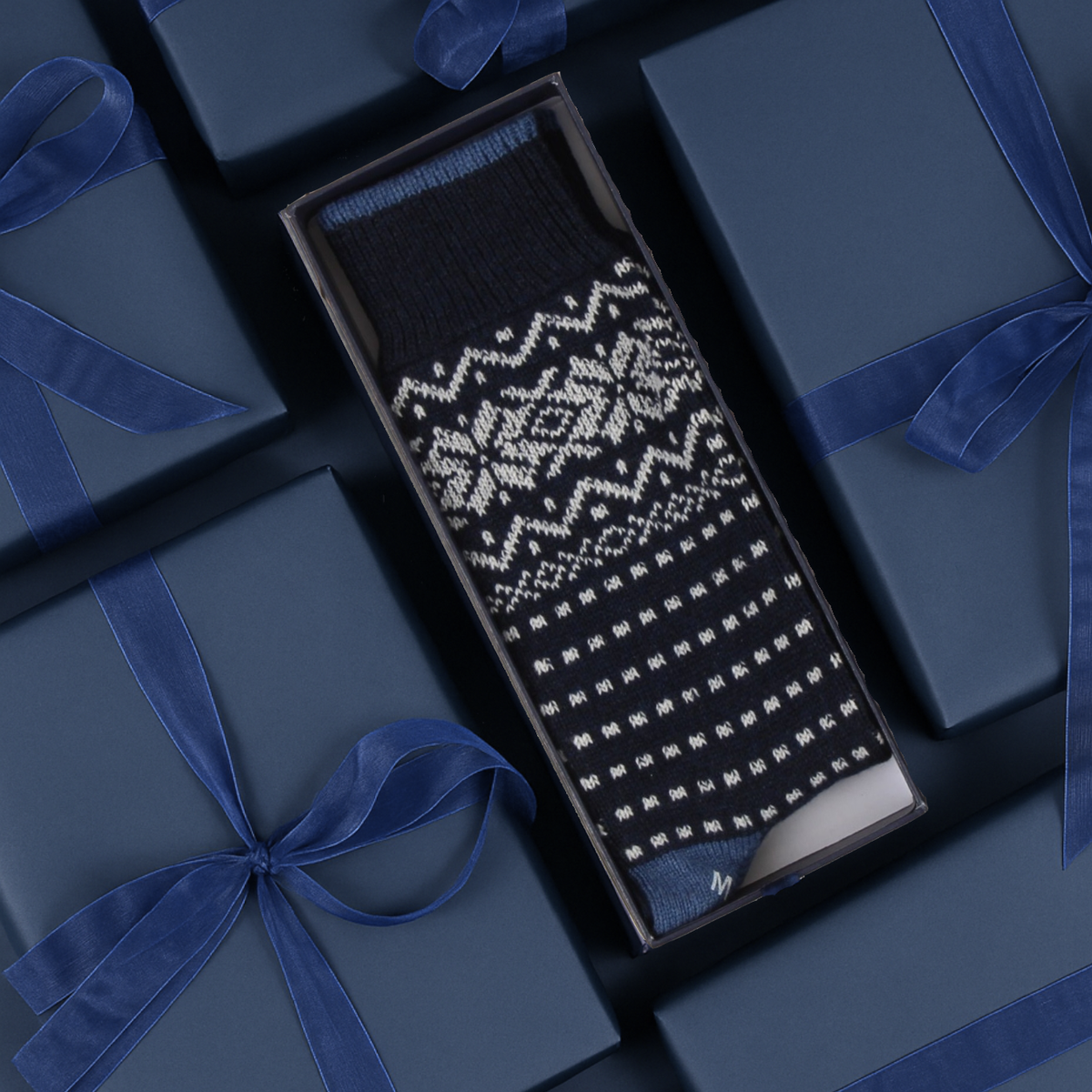 A single black knit sock with a white Fair Isle snowflake and geometric pattern, presented in an open dark gift box surrounded by navy blue gift boxes with blue ribbons.
