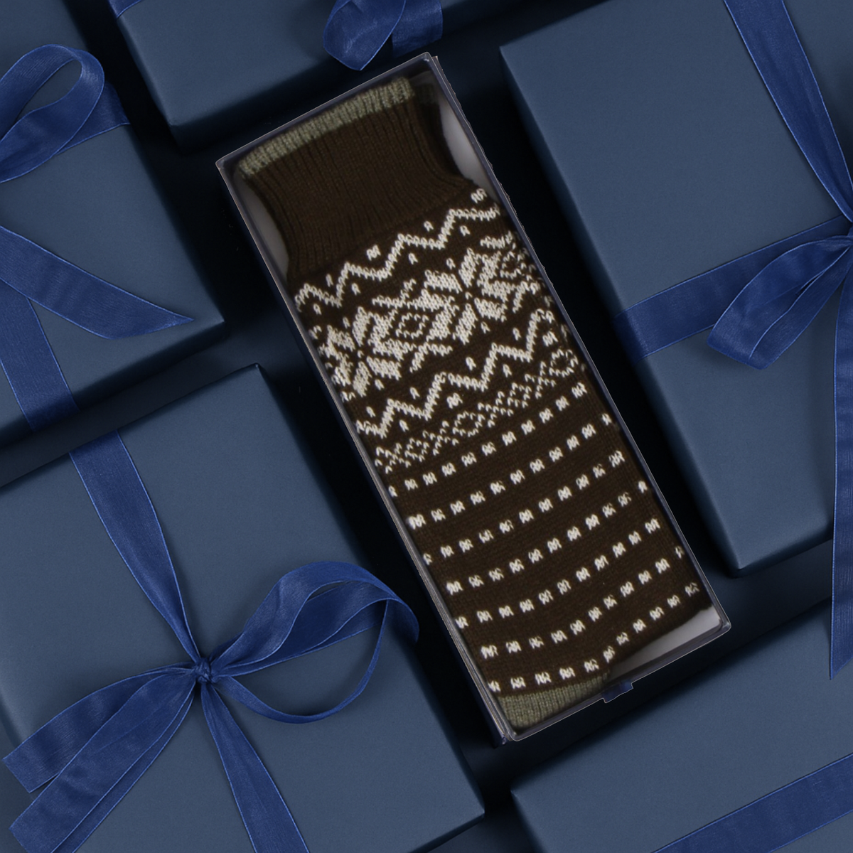 Socks with a white Fair Isle snowflake and geometric pattern, presented in an open dark gift box surrounded by navy blue gift boxes with blue ribbon.
