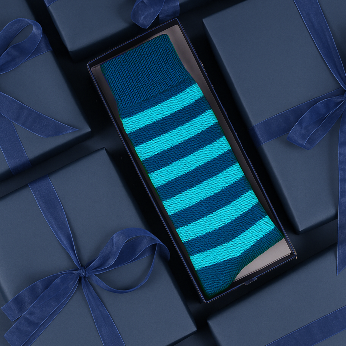 A single striped knit sock in a dark box, surrounded by navy gift boxes with blue ribbons. The sock has horizontal stripes in bright turquoise and dark teal-blue.