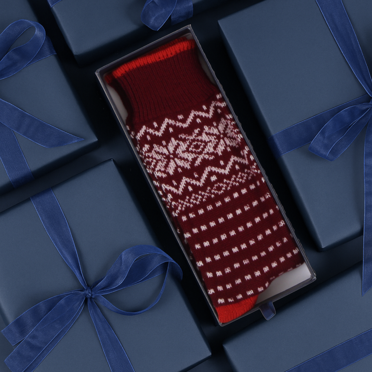Socks with a white Fair Isle snowflake and geometric pattern, presented in an open dark gift box surrounded by navy blue gift boxes with blue ribbon.