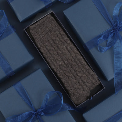 A pair of hand-knitted cable pattern cashmere socks presented in an open navy gift box with a socks pattern, surrounded by blue ribbon.