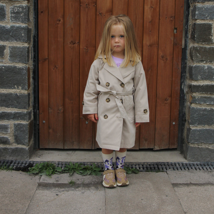 Young girl in a beige trench coat standing in front of a wooden door.