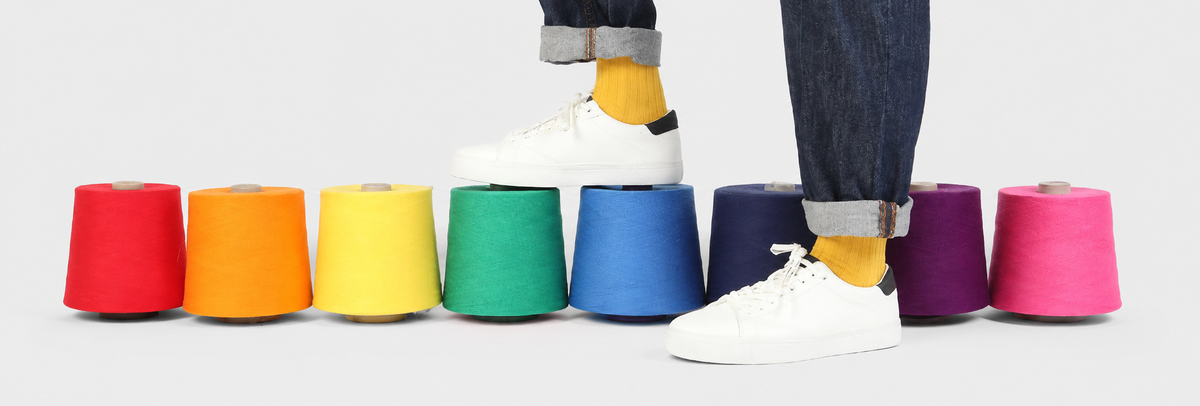 A person wearing white sneakers and dark jeans stands behind a row of colorful cone-shaped spools or color samples on a light gray background.