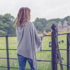 Woman in a gray poncho standing by a gate in a field with trees and buildings in the background
