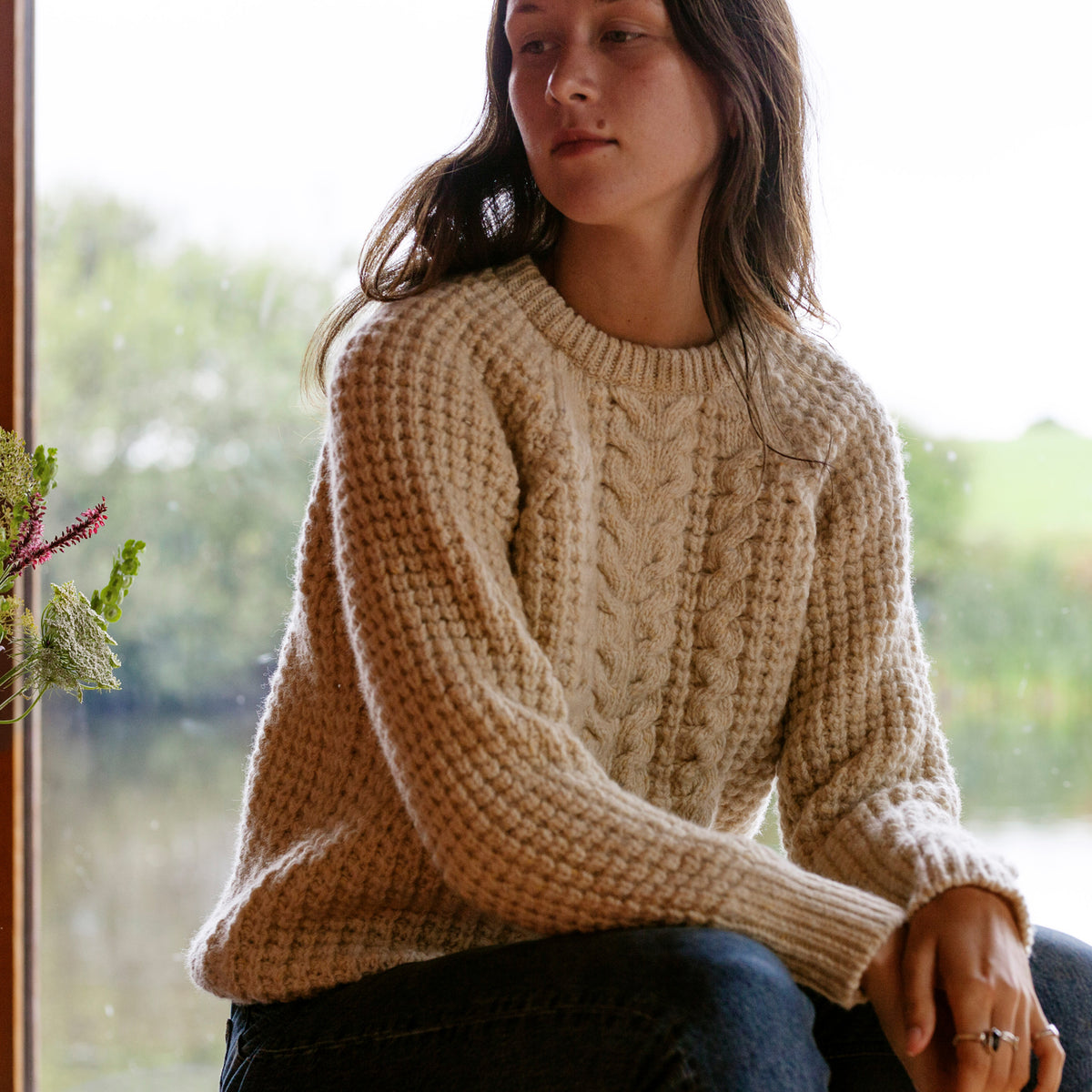 A woman sitting by a window wearing a beige chunky knit wool sweater with a round neckline and long sleeves.