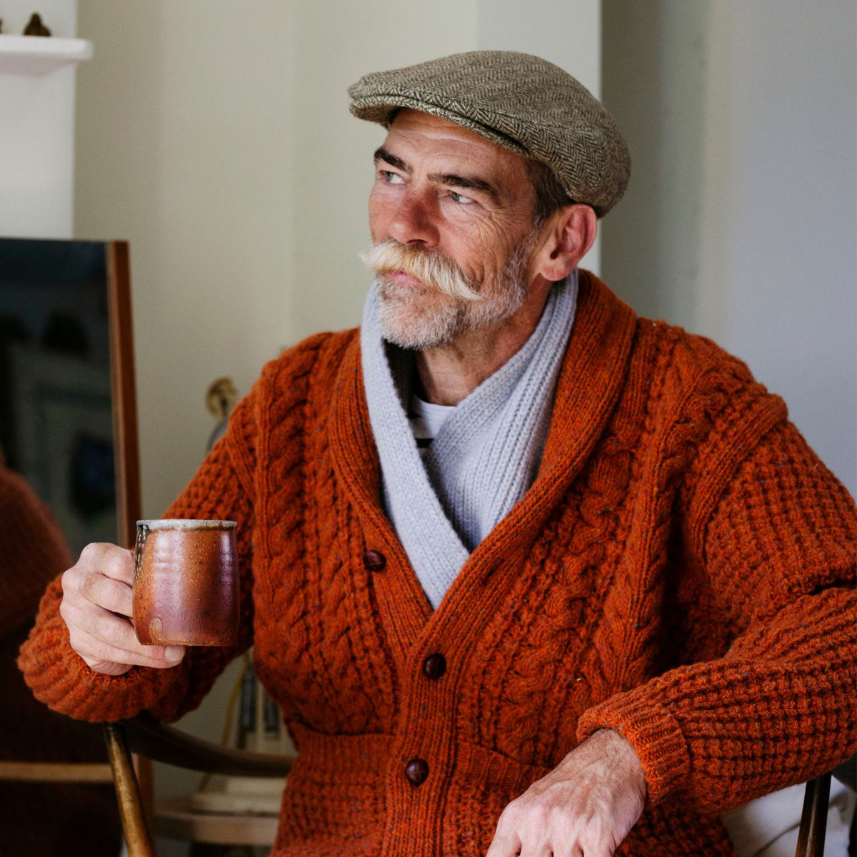 An older man with a beard and mustache wearing a brown cable knit cardigan over a grey sweater, paired with a beige flat cap, sitting and holding a copper mug.